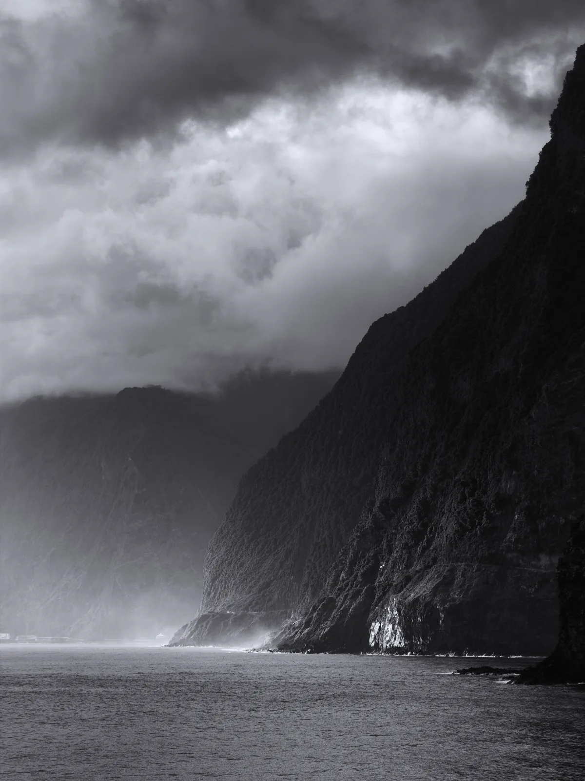 A dramatic cool black-and-white coastal cliff print with a dark cloud ceiling and luminous sea mist.