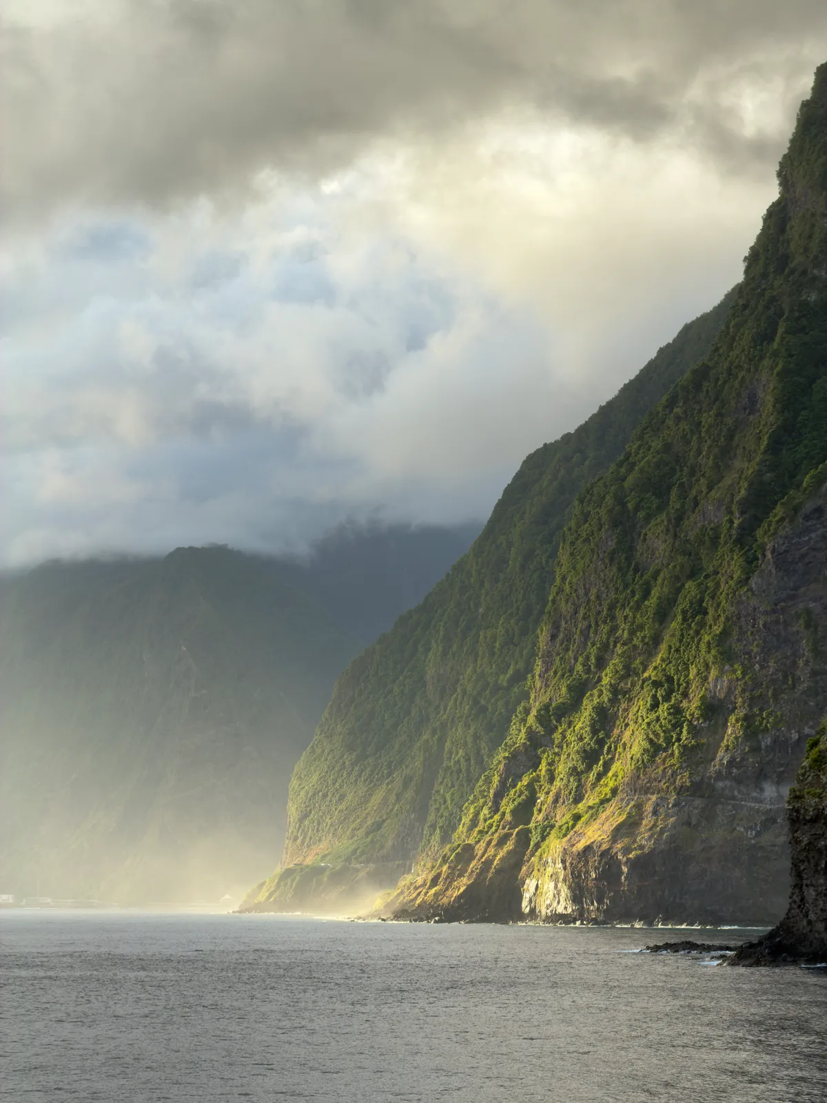 A color photograph of a steep coastal cliff beneath layered clouds and sea mist.